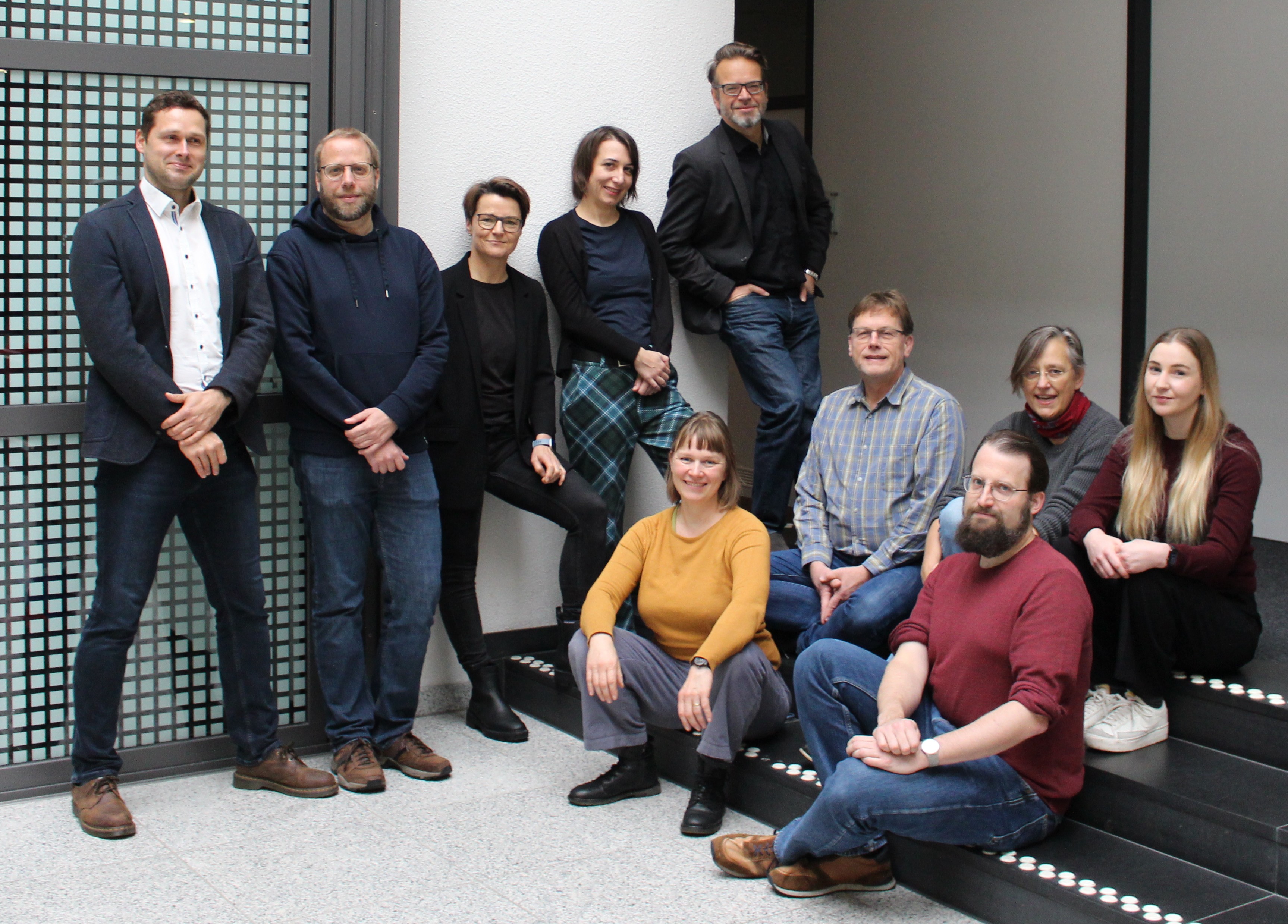Mehrere Personen stehen und sitzen in lockerer Gruppe auf einer Treppe und vor einer Wand.