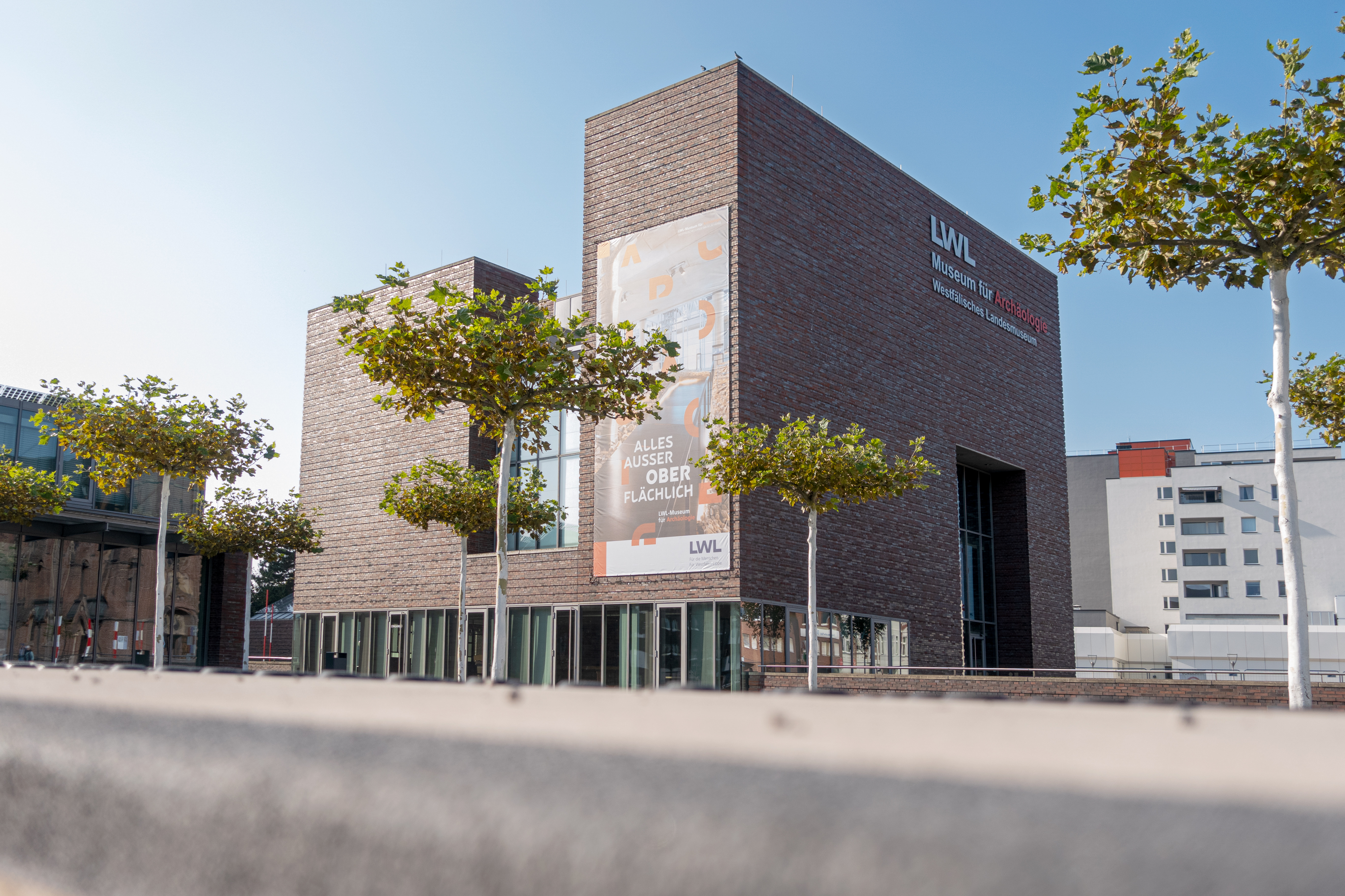 Modernes Backsteingebäude mit großen Fenstern und Banner mit Text 'ALLES AUSSER OBER FLÄCHLICH' vor blauem Himmel und Bäumen.
