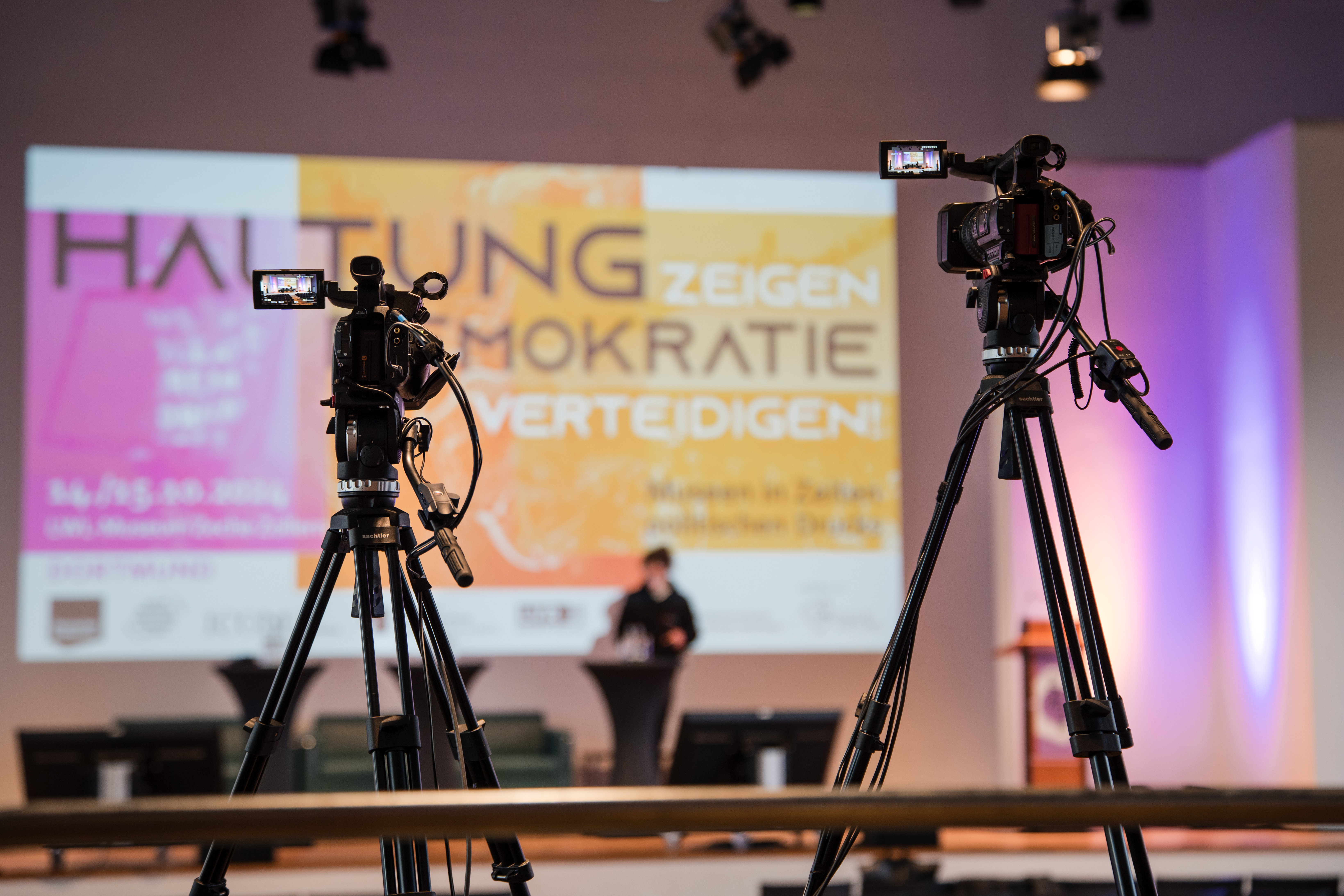 Zwei Kameras auf Stativen vor einer Bühne mit unscharfem Hintergrund und einer großen Projektionsfläche mit dem Text 'Haltung zeigen Demokratie verteidigen'