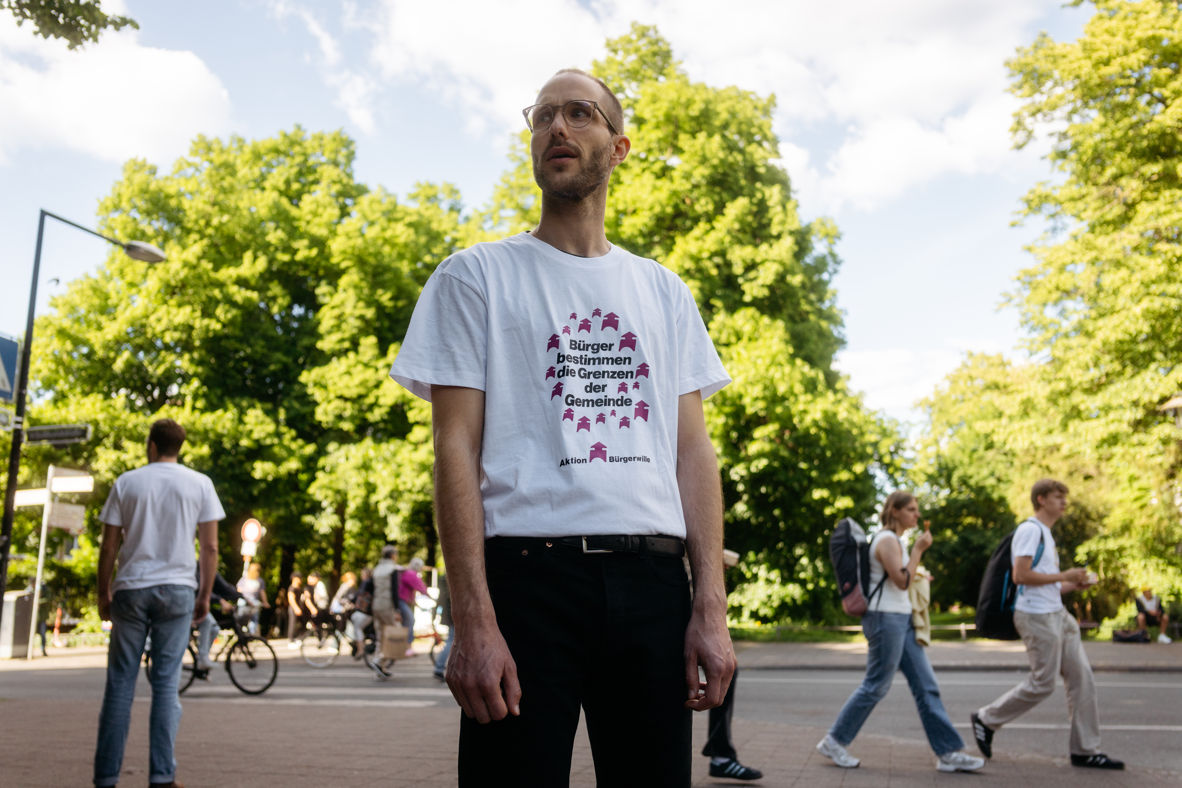 Mann mit weißem T-Shirt mit Aufdruck und schwarzer Hose steht auf einem Bürgersteig vor einer Straße, im Hintergrund mehrere Personen und grüne Bäume