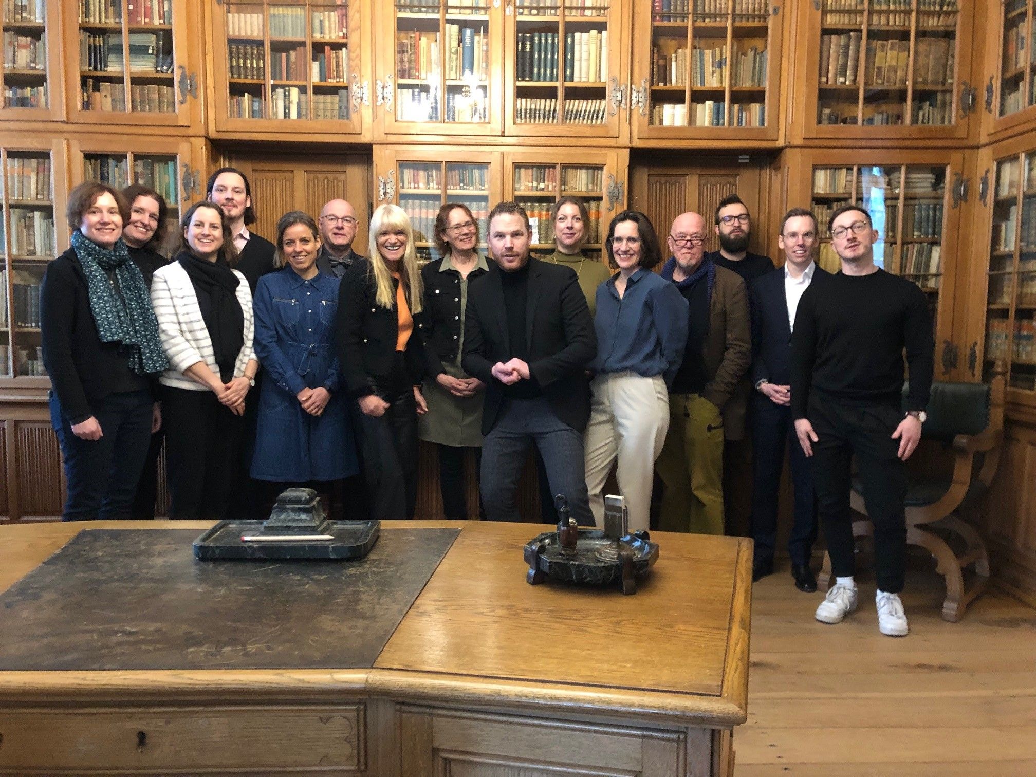 Gruppenfoto von etwa 15 Personen in einem historischen Raum mit hohen Holzregalen voller Bücher, vor einem großen Holztisch, alle in die Kamera blickend.