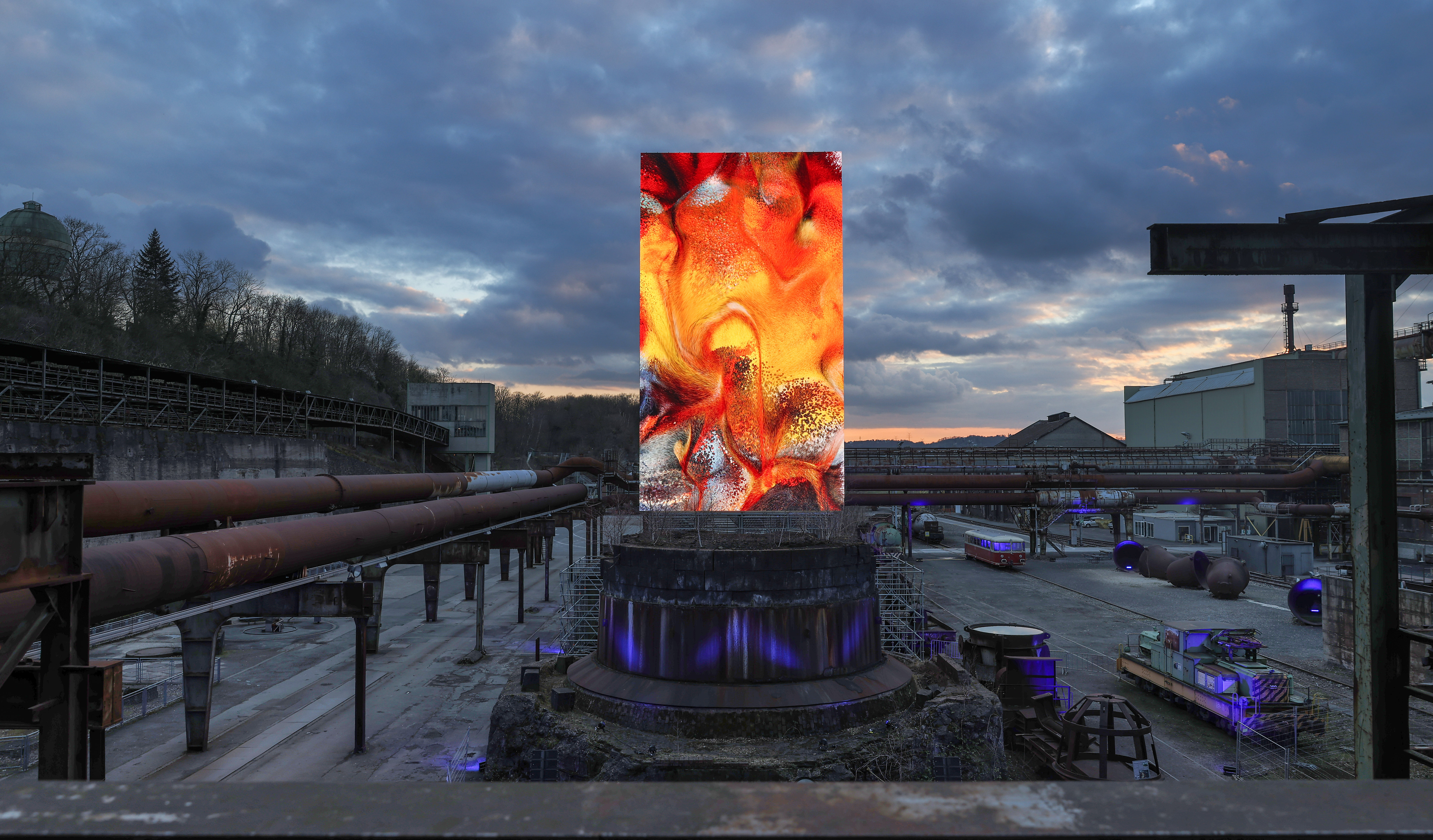 Monumentale digitale Datenskulptur von Refik Anadol mit rot-orangem, fließendem Muster auf einem runden Sockel im Industrieareal der Henrichshütte Hattingen bei Dämmerung.