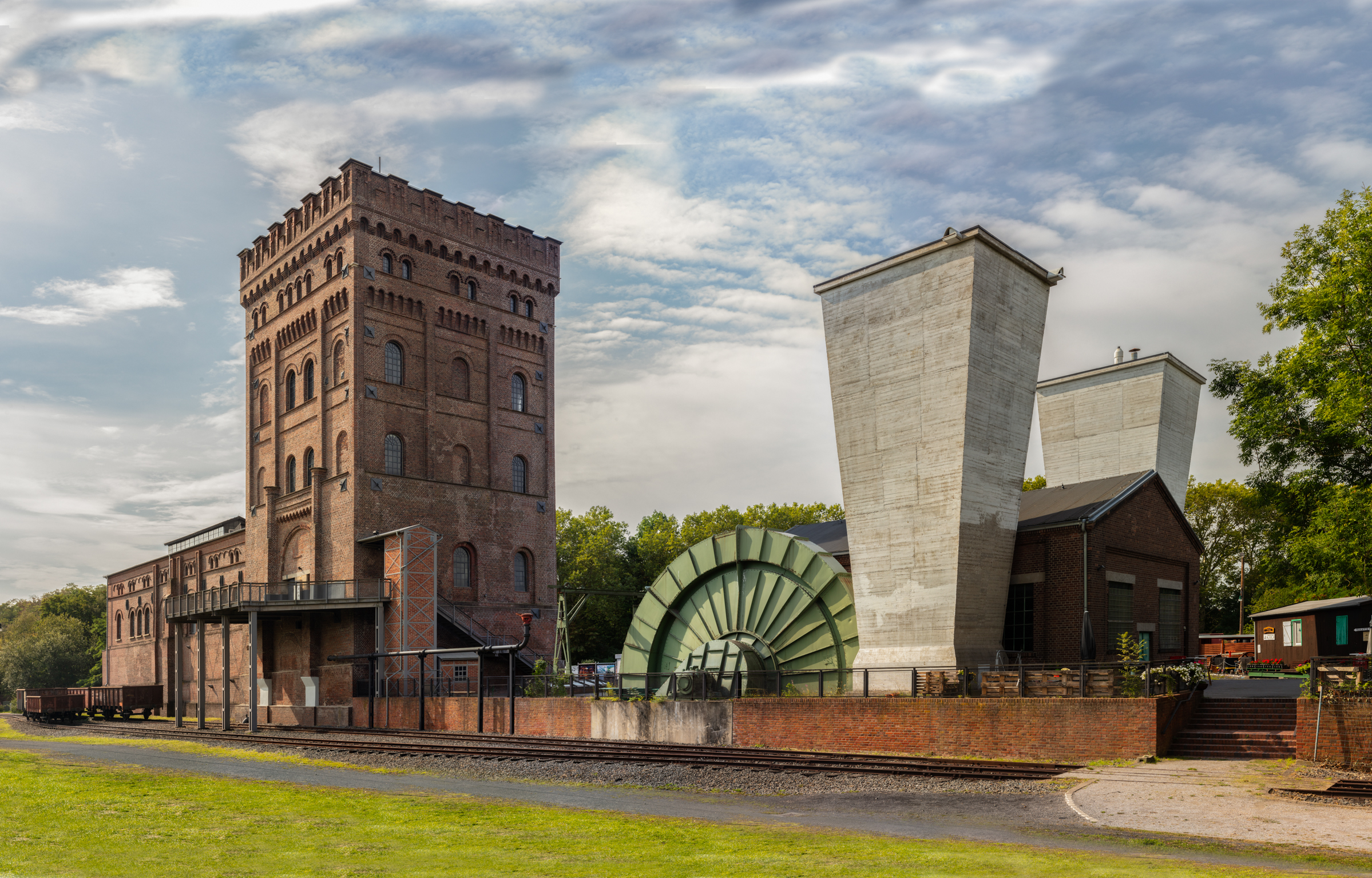 Industrieanlage mit rotem Backsteinturm, großem grünem Wasserrad und zwei hohen, trapezförmigen Betonschornsteinen unter bewölktem Himmel.