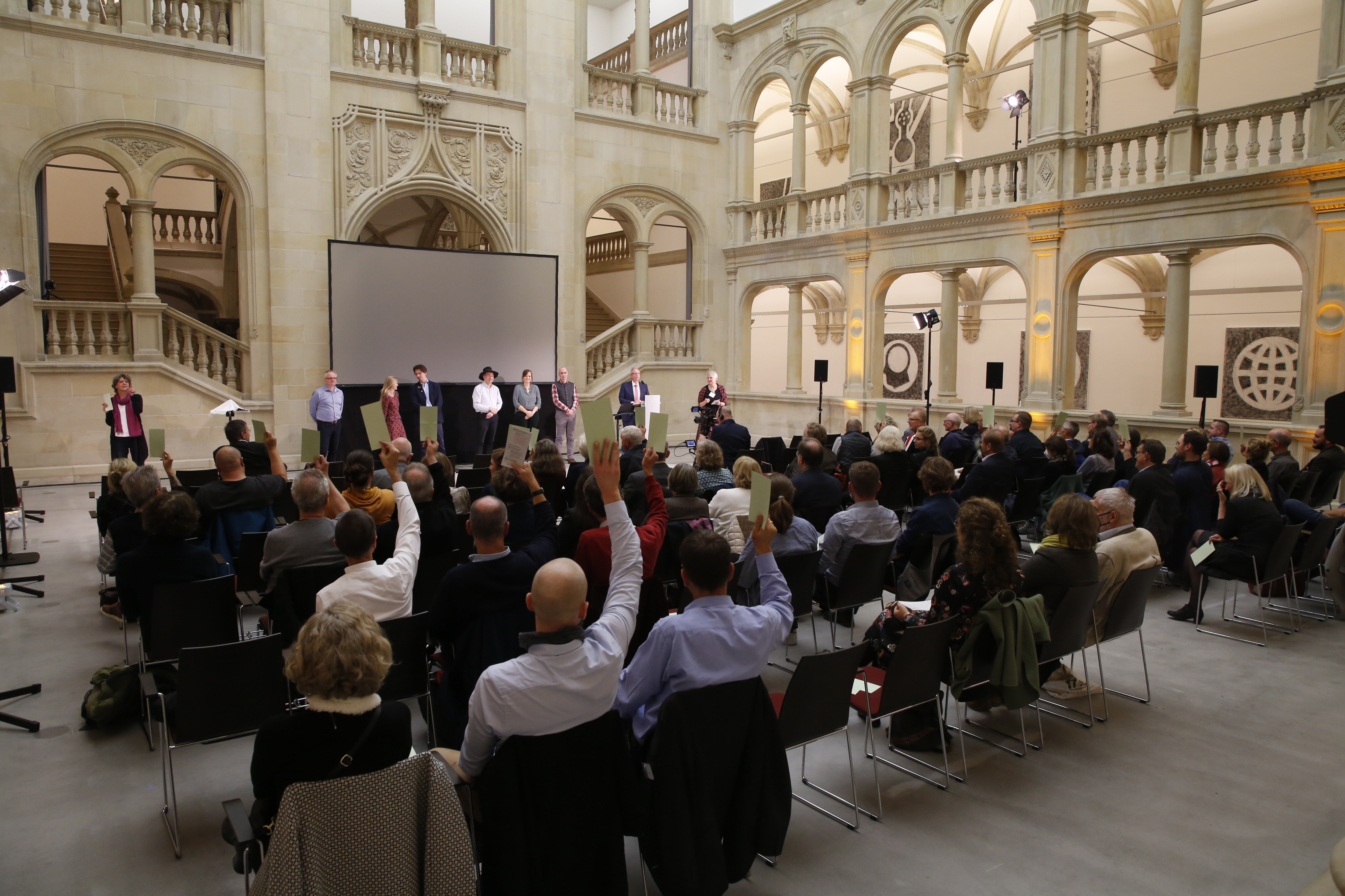 Mehrere Personen stehen vor einem Publikum in einem großen Saal mit hohen Fenstern und einer Treppe, einige Zuschauer heben die Hand zur Abstimmung.
