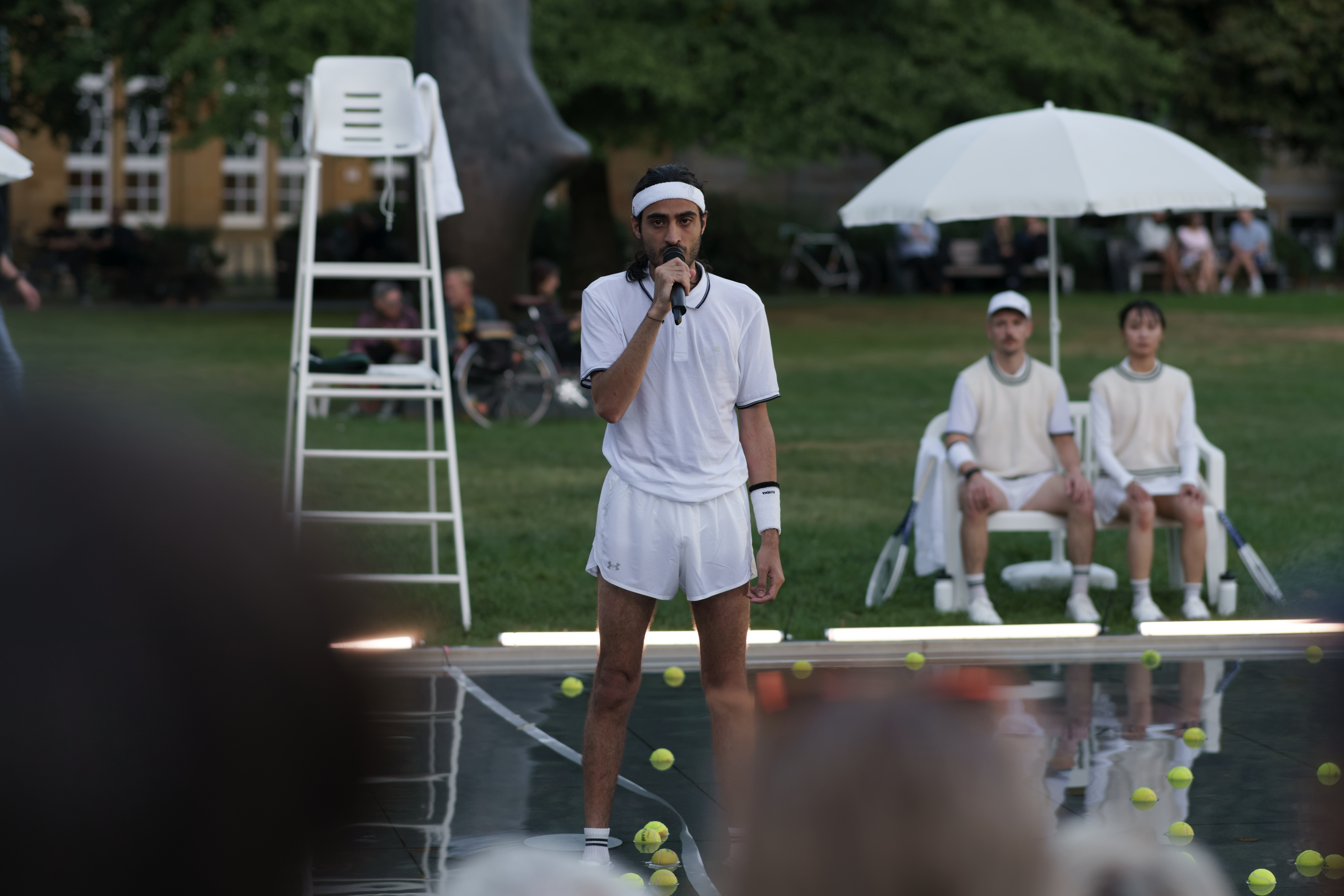 Ein Mann in weißer Tennisbekleidung steht auf einer Bühne im Freien und spricht in ein Mikrofon. Um ihn herum liegen zahlreiche Tennisbälle, während im Hintergrund zwei Personen unter einem Sonnenschirm sitzen und zuschauen.