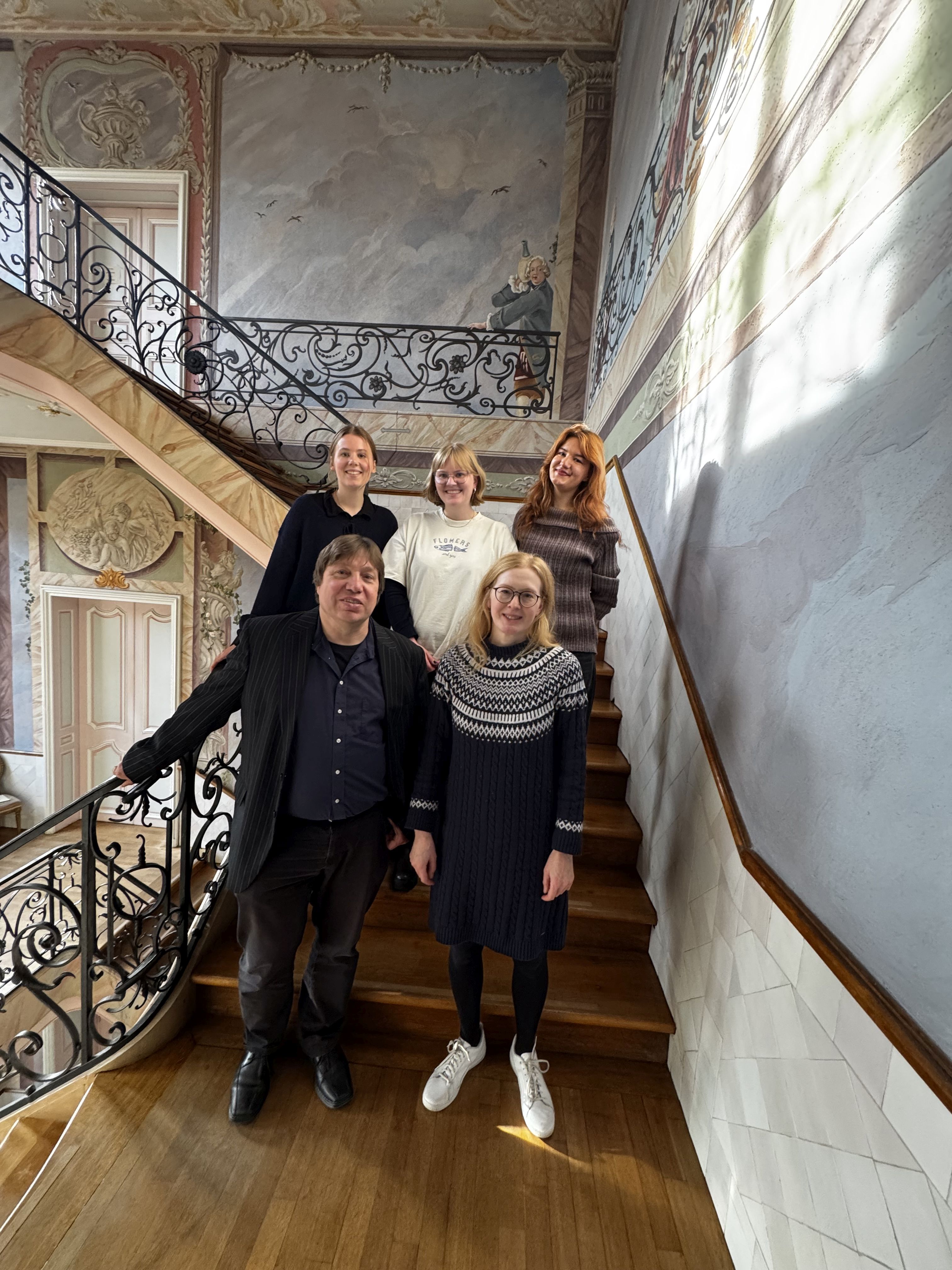 Fünf Personen stehen auf einer Treppe in einem historischen Gebäude mit verzierten Geländern und Wandmalereien.