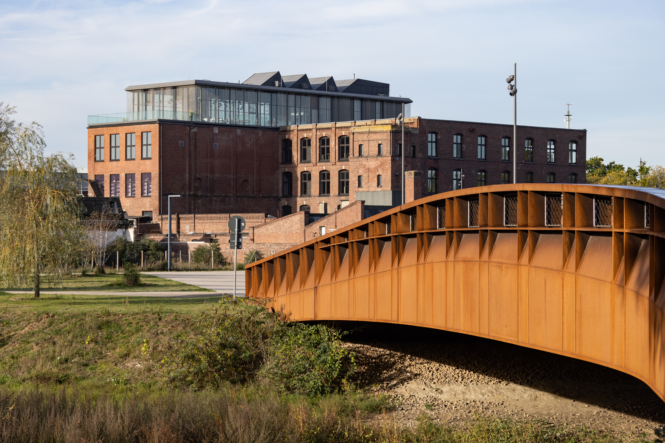 Blick auf altes Industriegebäude aus Backstein mit modernem Glasaufbau, im Vordergrund eine rostrote breite Brücke.