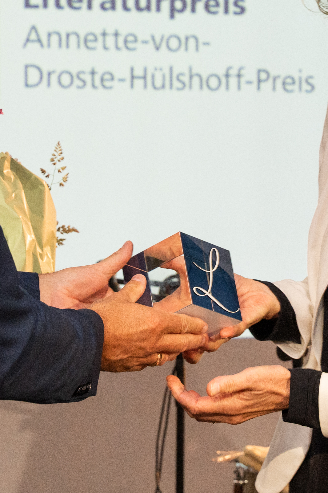 Zwei Hände überreichen eine geometrische Trophäe vor einer Bühne mit dem Schriftzug „Literaturpreis Annette-von-Droste-Hülshoff-Preis“.