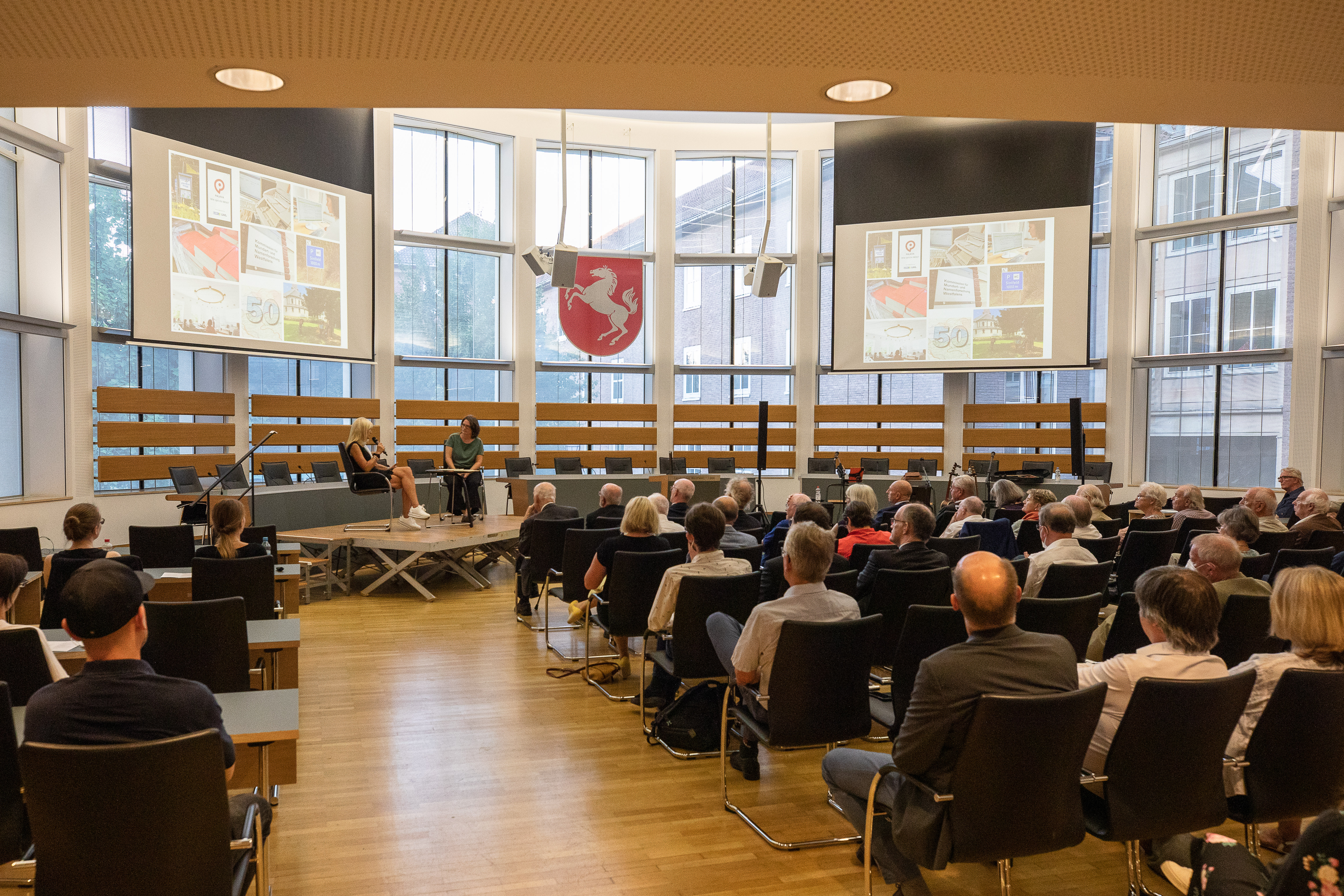 Publikum sitzt in einem Saal mit großen Fenstern vor einer Bühne, auf der zwei Personen an Tischen mit Mikrofonen sitzen, darüber ein rotes Wappen mit weißem Pferd.