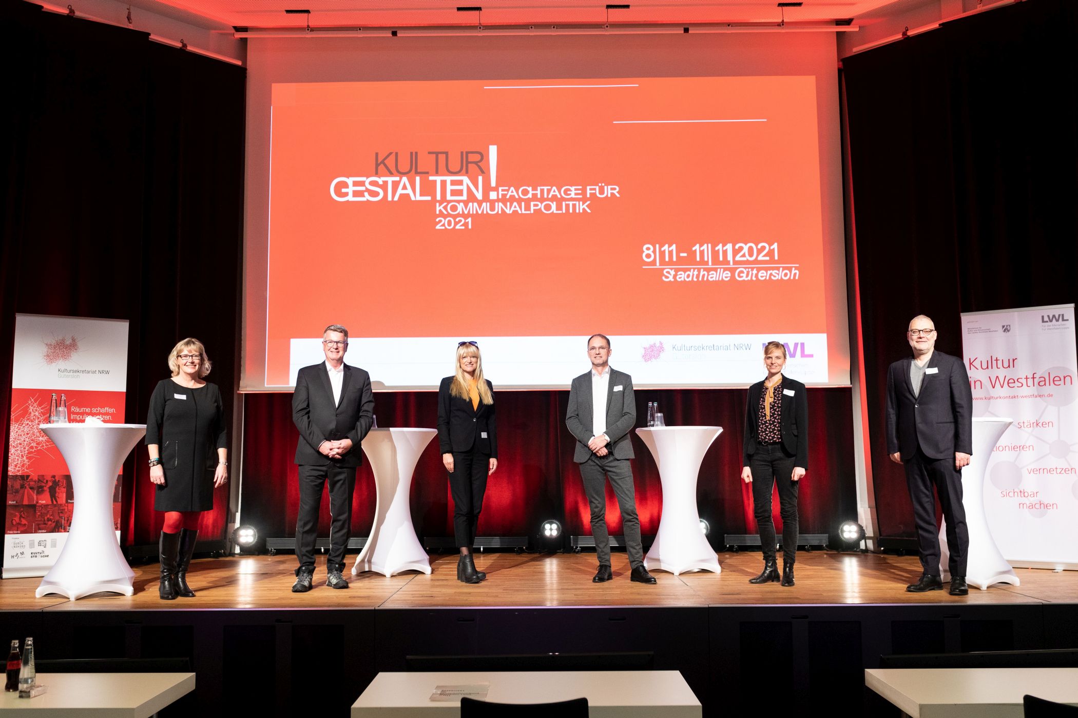 Sechs Personen stehen mit Abstand auf einer Bühne vor einem roten Präsentationsbildschirm und weißen Stehtischen in einem Konferenzsaal