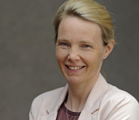 Porträt einer lächelnden Frau mit blondem Haar, die einen hellen Blazer und eine dunkle Kette trägt, vor neutralem Hintergrund