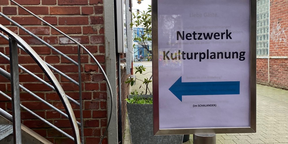 Schild mit der Aufschrift 'Netzwerk Kulturplanung' und einem Pfeil nach links neben einer Metalltreppe und einem roten Absperrband in einem Innenhof