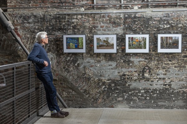 Person lehnt an einem Geländer und betrachtet vier gerahmte Fotografien an einer rauen Steinwand in einem Ausstellungsraum.