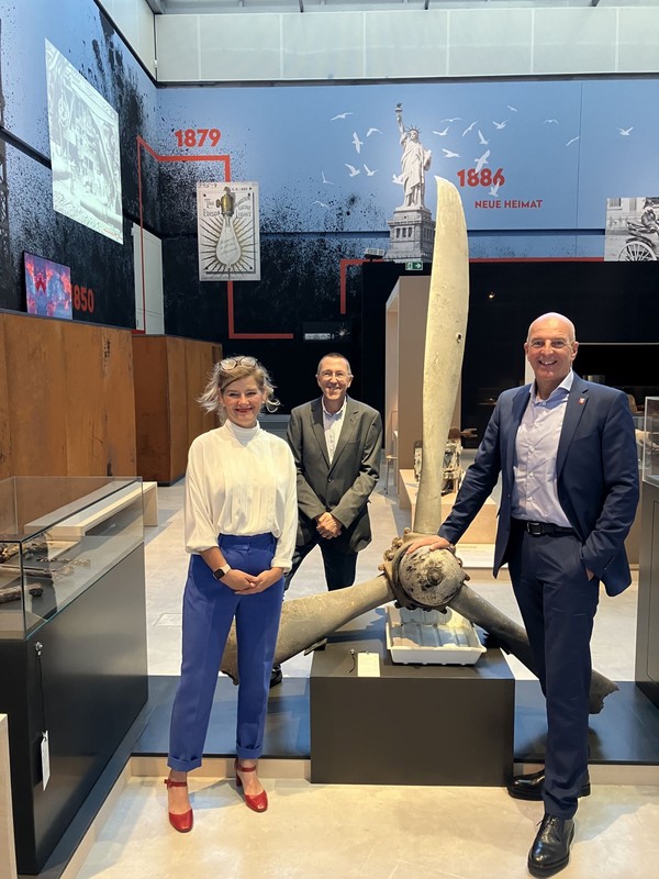 Drei Personen in formeller Kleidung stehen in einem Museum vor einem großen Propeller, im Hintergrund eine Wand mit einem Zeitstrahl.