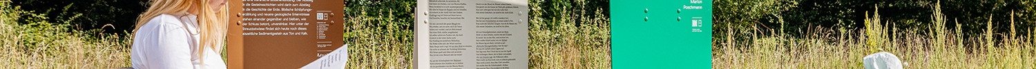 Eine Person sitzt draußen auf einer Holzbank vor Outdoor-Ausstellungsstelen und einem Tisch mit Ausstellungsobjekt