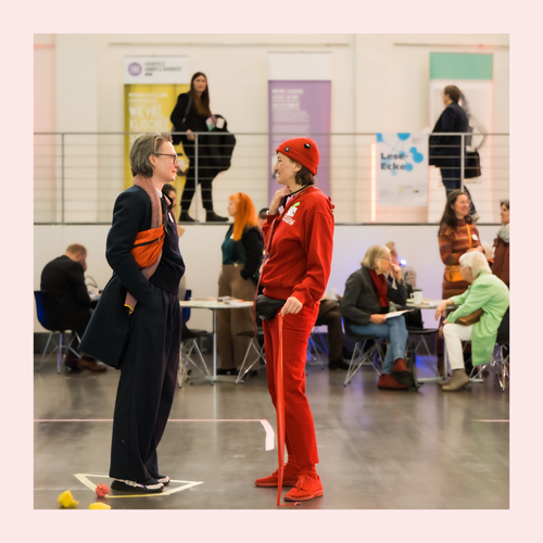 Zwei Personen stehen sich in einem großen Raum gegenüber, im Hintergrund sitzen und stehen weitere Menschen an Tischen und auf einer Galerie.