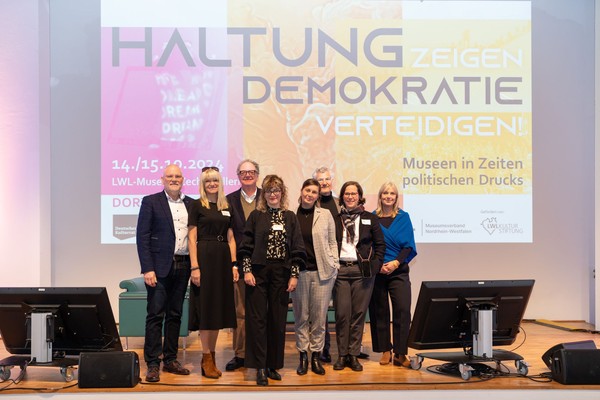 Sieben Personen stehen auf einer Bühne vor einer Leinwand mit der Projektion 'HALTUNG ZEIGEN DEMOKRATIE VERTEIDIGEN!' und weiteren Texten zu einer Tagung im LWL-Museum.