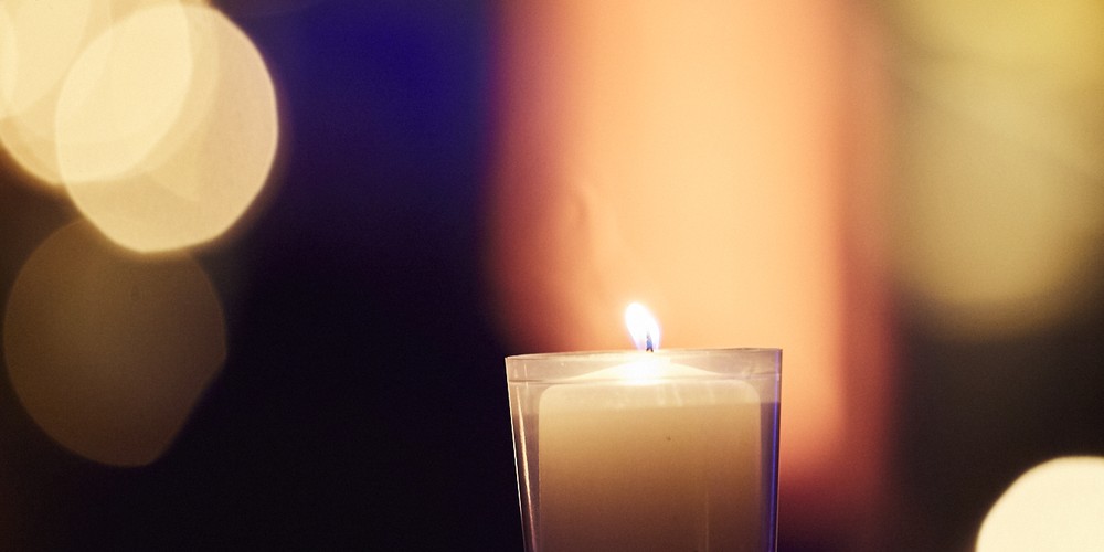 Brennende Kerze in durchsichtigem Glas vor unscharfem, farbigem Hintergrund mit Lichtpunkten