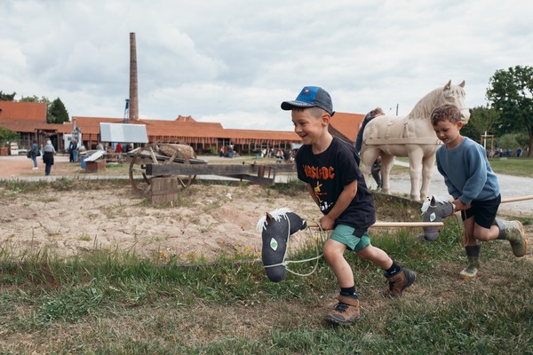 Zwei Kinder spielen auf einem Sandplatz mit Steckenpferden vor einer Industriegebäudekulisse.