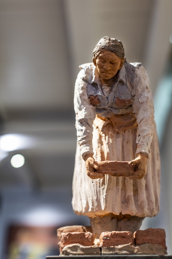 Figur einer Frau in traditioneller Kleidung, die Ziegelsteine hält, vor unscharfem Hintergrund.