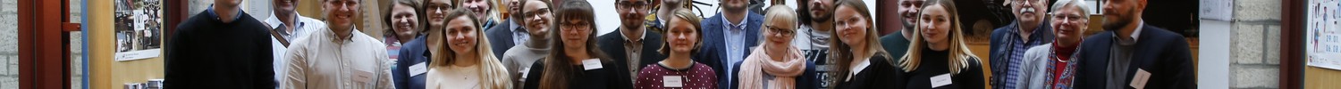 Gruppe von Personen steht in einem Raum mit großem runden Uhrwerk an der Decke und großen Fenstern im Hintergrund.