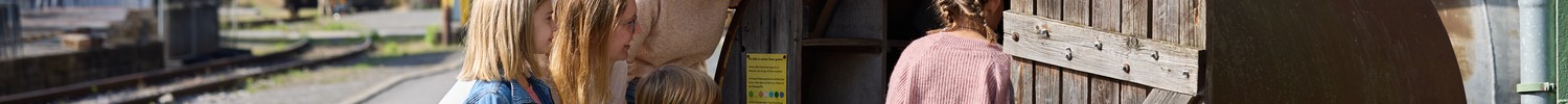 Gruppe von Kindern und eine erwachsene Person, die in ein rostiges Gefäß mit offener Holztür schauen, davor eine blaue Rattenfigur an der Holztür.