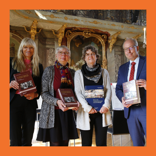Vier Personen stehen nebeneinander in einem prunkvollen Raum und halten jeweils ein Buch in den Händen. Foto ist mit einem orangenem Rahmen gerahmt.