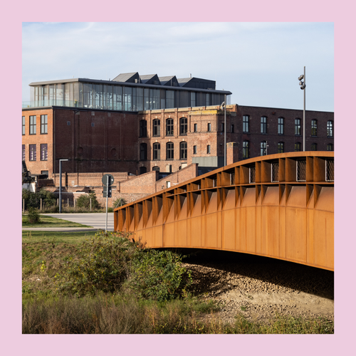 Blick von der Weberei auf das Backsteingebäude der Spinnerei, im Vordergrund eine Brücke über den Fluss, Bild eingefasst von einem rosa Rahmen