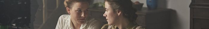 Zwei Personen sitzen an einem Tisch in einem Raum mit rustikaler Einrichtung und schälen Kartoffeln. Die Szene wird von einer Filmkamera beleuchtet.
