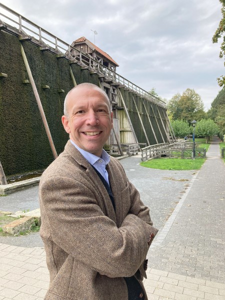 Mann mit braunem Strickjacke und blauem Hemd steht lächelnd mit verschränkten Armen vor einer großen Holzkonstruktion und einem Weg mit Bäumen