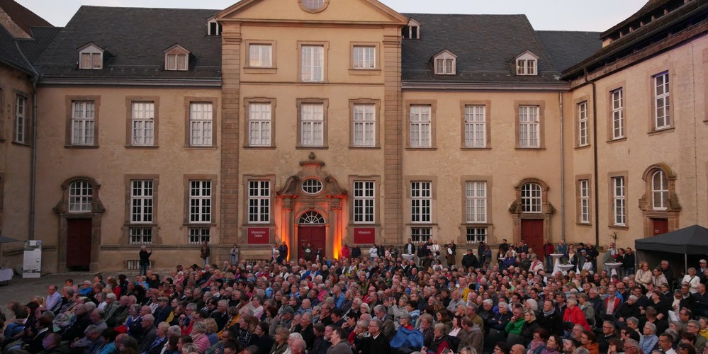 Große Menschenmenge sitzt vor einem historischen Gebäude mit vielen Fenstern und einem Uhrturm auf dem Dach