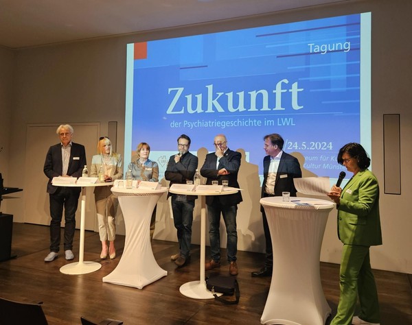 Sieben Personen stehen an drei hohen Tischen auf einer Bühne vor einer Leinwand mit der Projektion 'Tagung Zukunft der Psychiatriegeschichte im LWL'.