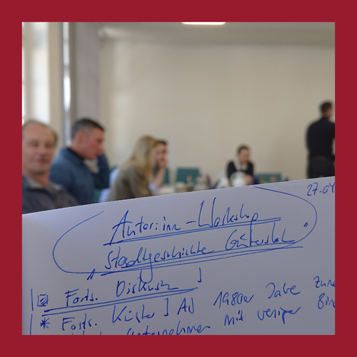 Handschriftliche Notizen auf einem Blatt Papier im Vordergrund, im Hintergrund unscharfe Personen in einem Raum sitzend