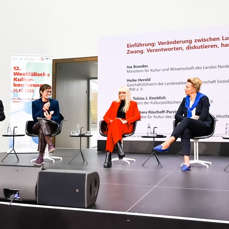 Drei Personen sitzen auf Stühlen auf einer Bühne vor einer Wand mit Text und einem Banner der 12. Westfälischen Kulturkonferenz