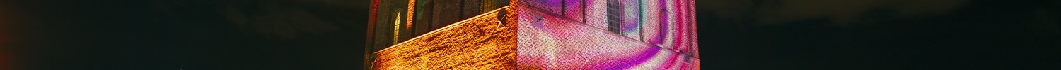 Historischer Backsteinturm bei Nacht mit farbiger Lichtprojektion und Menschen davor.