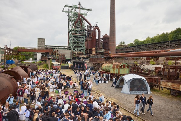 Viele junge Menschen unterhalten sich in Grüppchen auf einem Industriegelände. Im Hintergrund eine Bühne vor dem Hochofen.