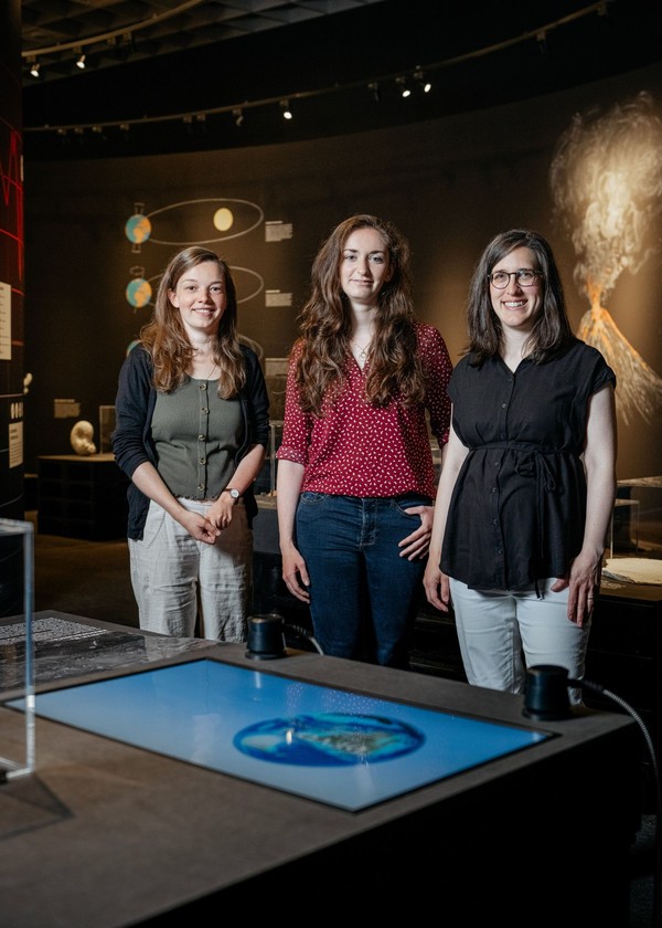 Drei Personen stehen nebeneinander in einem Museum vor einer interaktiven Ausstellung mit einem Bildschirm und wissenschaftlichen Grafiken an der Wand