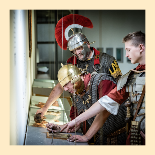 Foto eingefasst in einen gelben Rahmen. Foto von römischen Legionären, die auf Wachstafel schreiben.
