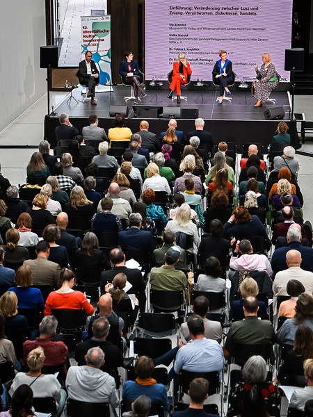 Publikum sitzt vor einer Bühne mit fünf sitzenden Podiumsteilnehmern und einer großen Leinwand mit Text im Hintergrund
