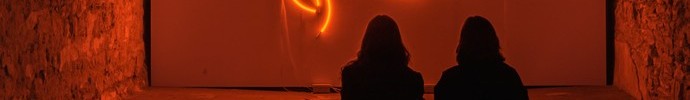 Zwei Personen sitzen vor einer leuchtenden orangefarbenen Neoninstallation in einem dunklen, gewölbten Raum mit Backsteinwänden.