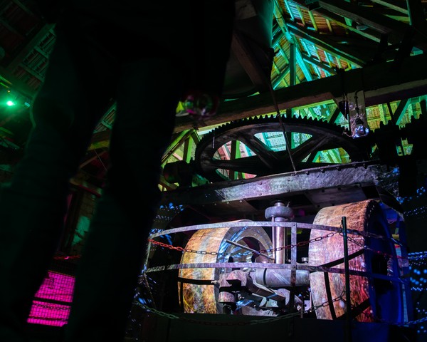 Lichtinstallation: Erleuchtete große Zahnräder und Maschinenteile unter einem grün beleuchteten Dach