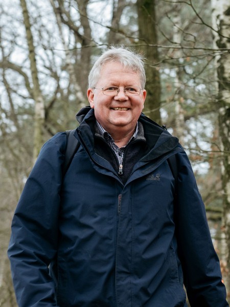 Älterer Mann mit Brille und dunkelblauer Jacke geht lächelnd auf einem Waldweg zwischen kahlen Bäumen