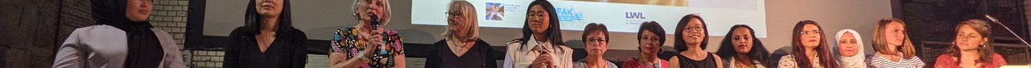 Gruppe von Frauen steht vor einer Leinwand mit dem Text 'Ich nenne es hier auch Zuhause' in einem großen Raum mit Backsteinwänden