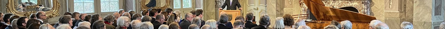 Festlich gekleidete Menschen sitzen in einem prunkvollen Saal mit Kronleuchter und großem Gemälde, eine Person spricht an einem Rednerpult.