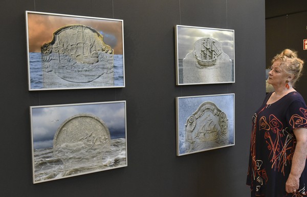 Vier gerahmte Fotografien von steinernen Reliefs mit Schiffsmotiven an einer schwarzen Wand, daneben eine Frau mit gemustertem Kleid