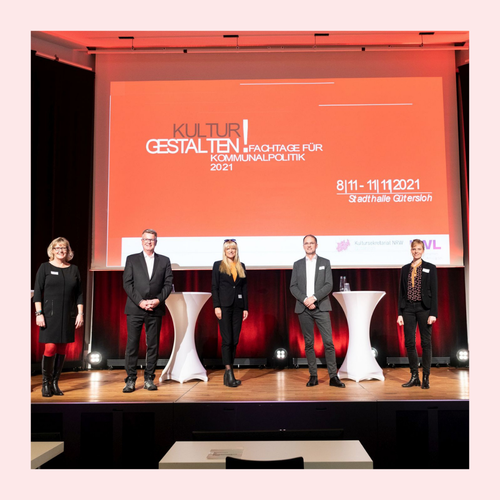 Sechs Personen stehen mit Abstand auf einer Bühne vor einem roten Präsentationsbildschirm und weißen Stehtischen in einem Konferenzsaal. Foto eingerahmt von rosa Rahmen.