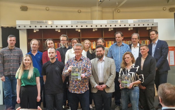 Eine Gruppe von Menschen vor einem Bücherregal, zwei Personen halten Bücher in die Kamera.