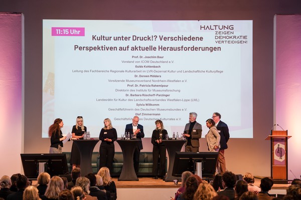Sieben Personen stehen auf einer Bühne vor einem großen Bildschirm mit dem Titel der Veranstaltung und diskutieren vor einem Publikum.
