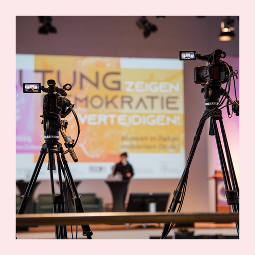 Zwei Kameras auf Stativen vor einer Bühne mit unscharfem Hintergrund und einer großen Projektionsfläche mit dem Text 'Haltung zeigen Demokratie verteidigen'
