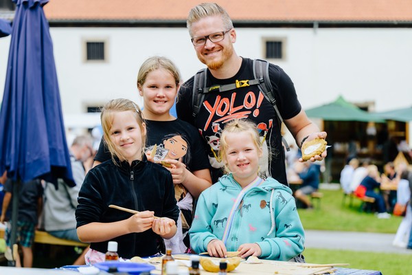 Familie mit drei Kindern steht an einem Tisch im Freien und hält Bastelmaterial in den Händen